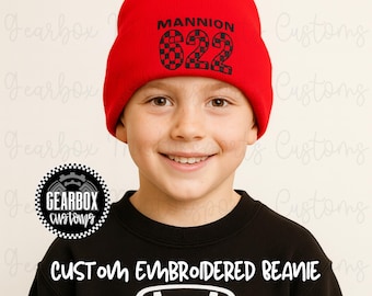 May include: A red beanie with the name "MANNION" and the number "622" in a checkered pattern. The beanie is embroidered and worn by a person. The text "CUSTOM EMBROIDERED BEANIE" is below the person.