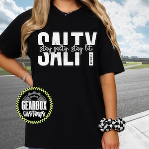 Puede incluir: Camiseta negra con las palabras blancas "Salty Salt". La frase "stay salty, stay lit" está encima. El texto "Matthew 5:13" está en un rectángulo blanco. Un logotipo de Gearbox Customs está en la parte inferior izquierda. Ropa casual.