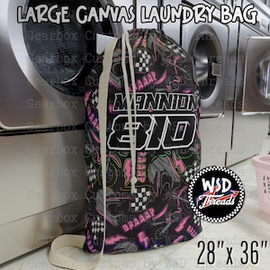 May include: Large canvas laundry bag with a black background and a colorful design featuring racing-themed graphics. The bag has a drawstring closure and a beige strap. The bag measures 28 inches by 36 inches.