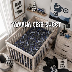 Könnte beinhalten: Ein Holzbettchen mit einem Yamaha-Bettlaken. Das Laken hat ein Muster aus Yamaha-Logos, Schachflaggen und blauen Akzenten auf dunklem Hintergrund. Der Raum hat ein "Rad Dude"-Schild, einen Motorrad-Druck und ein Spielzeugmotorrad.