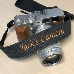 May include: A silver digital camera with a brown leather grip and a gray strap that reads "Jack's Camera" in orange embroidery. A silver lens is in front of the camera.