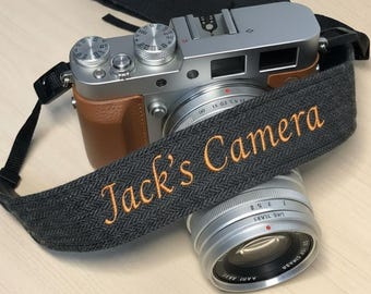 Puede incluir: Cámara digital plateada con empuñadura de cuero marrón y correa gris con bordado naranja "Jack's Camera". Una lente plateada está frente a la cámara.