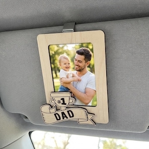 May include: A light-coloured wooden car visor clip frame featuring a rectangular photo of a person holding a child. The frame includes a decorative wooden cutout at the bottom shaped like a trophy with the text #1 DAD engraved on a banner.