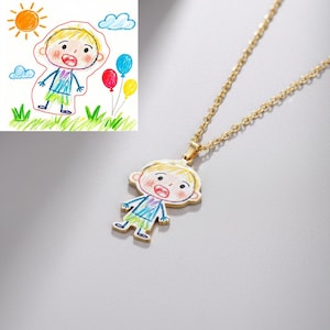 Op de afbeelding: Een goudkleurige ketting met een hanger in de vorm van een kindertekening. De tekening toont een persoon met een blauw shirt, groene shorts en een verbaasde uitdrukking. De ketting ligt op een grijs oppervlak, met een tekening van een zon, wolken en ballonnen op de achtergrond.