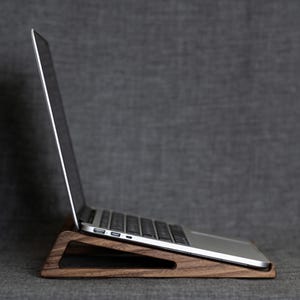 May include: A laptop computer rests on a wooden stand. The stand is made of dark brown wood and has a triangular design with a cutout in the center. The laptop is silver and black, with the screen open.