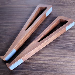 May include: Two wooden laptop stands with a triangular design. The stands are made of light brown wood and feature a light blue pad on the bottom. The stands are designed to elevate a laptop for better ergonomics.