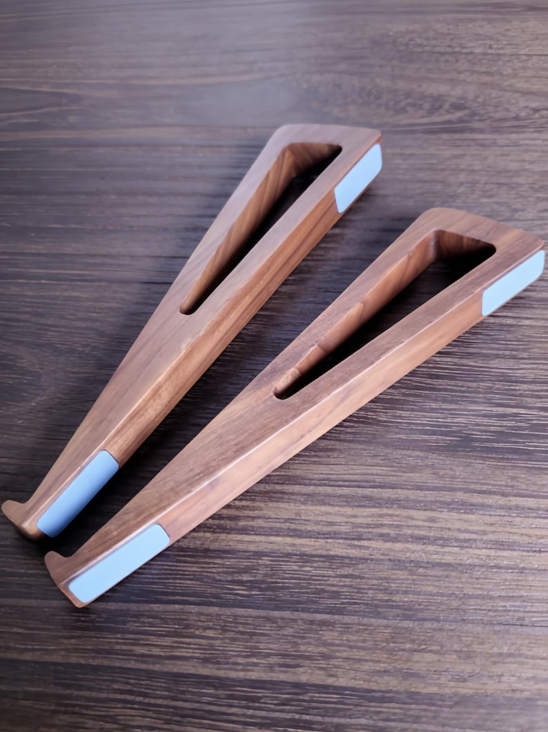 May include: Two wooden laptop stands with a triangular design. The stands are made of light brown wood and feature a light blue pad on the bottom. The stands are designed to elevate a laptop for better ergonomics.