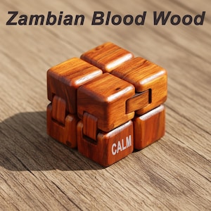 Puede incluir: Un cubo fidget de madera de Zambian Blood Wood. El cubo es de color marr&oacute;n c&aacute;lido con vetas de madera visibles. La palabra "CALM" est&aacute; impresa en una de las caras del cubo. El texto "Zambian Blood Wood" est&aacute; en la parte superior de la imagen.