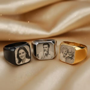 May include: Three signet rings with engraved photos. One is black, one silver, and one gold. Each ring has a square face with a black and white image of a person or couple. The rings are displayed on a gold fabric.