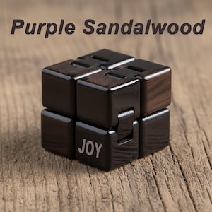 Puede incluir: Un juguete cubo fidget de madera de s&aacute;ndalo p&uacute;rpura. El cubo es de color marr&oacute;n oscuro con vetas de madera visibles y la palabra "JOY" est&aacute; impresa en uno de los bloques. El texto "Purple Sandalwood" est&aacute; encima del cubo.