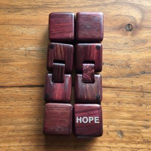 Puede incluir: Un juguete de madera para manipular de color rojo oscuro, con una serie de bloques cuadrados conectados. La palabra "HOPE" est&aacute; impresa en blanco en uno de los bloques. El juguete est&aacute; dise&ntilde;ado para ser manipulado y jugado.