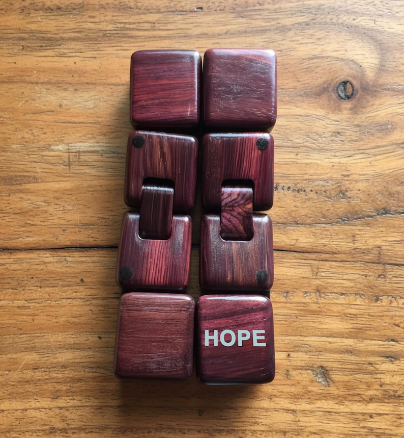 Puede incluir: Un juguete de madera para manipular de color rojo oscuro, con una serie de bloques cuadrados conectados. La palabra "HOPE" est&aacute; impresa en blanco en uno de los bloques. El juguete est&aacute; dise&ntilde;ado para ser manipulado y jugado.