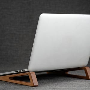 May include: A wooden laptop stand with a white laptop resting on it. The stand is made of light brown wood and has a triangular design. The laptop is white and has a silver edge.