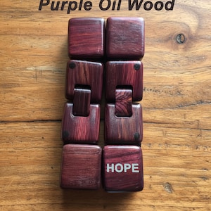 Puede incluir: Una pulsera de madera hecha de bloques cuadrados e interconectados de madera de aceite p&uacute;rpura. La palabra "HOPE" est&aacute; impresa en blanco en uno de los bloques. El texto "Purple Oil Wood" est&aacute; en la parte superior de la imagen.