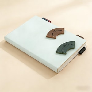 May include: A pale green notebook with brown and green leather page markers. The markers are semi-circular and have the words "Turn the page" printed on them. The notebook has several coloured page markers.