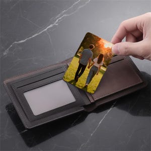 May include: A bi-fold wallet in shades of grey and brown, with a clear ID window. A rectangular card with a photo of a couple walking in a field is being held above the wallet.