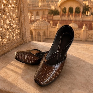 May include: A pair of dark brown slip-on shoes featuring a pointed toe and intricate metallic stitching patterns across the upper surface. The shoes have a dark interior lining and a flat sole, showcasing a detailed design with visible needlework.