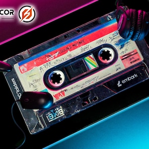 May include: A gaming mousepad featuring a retro cassette tape design with the text "ALGUNS MIXTAPE". A black controller, mouse, and headphones are also visible. The mousepad has a black background with pink and blue neon accents.