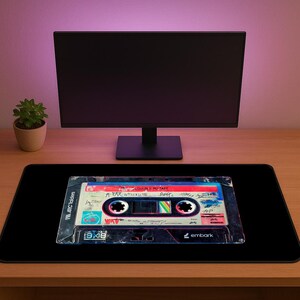 May include: Black rectangular mousepad featuring a vintage cassette tape design. The cassette tape has a colorful label with handwritten text and a retro aesthetic. The mousepad is on a wooden desk with a computer monitor and a small potted plant in the background.