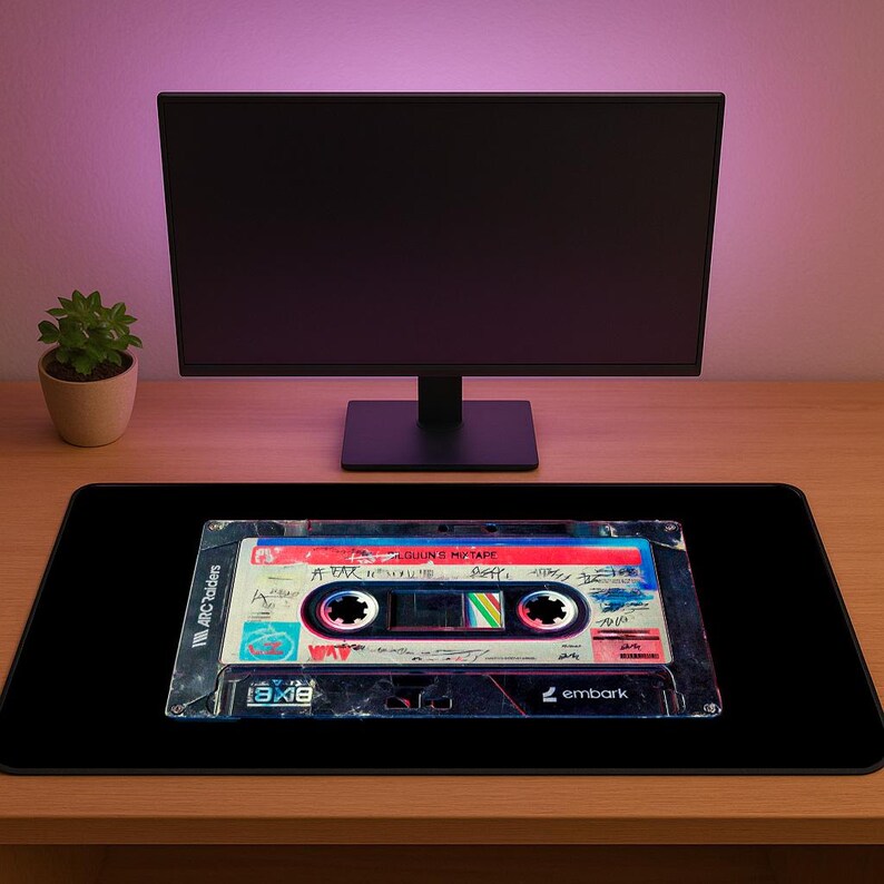 May include: Black rectangular mousepad featuring a vintage cassette tape design. The cassette tape has a colorful label with handwritten text and a retro aesthetic. The mousepad is on a wooden desk with a computer monitor and a small potted plant in the background.