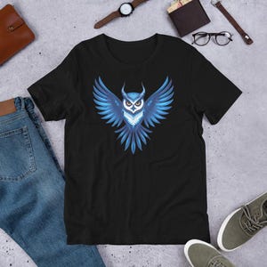 以下が含まれることがあります： 黒いTシャツ。青いフクロウのデザインで、翼を広げている。フクロウのデザインは、白い胸と黄色の目があり、グラデーション効果がある。柔らかい素材で作られたTシャツ。