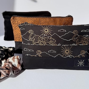 May include: Three rectangular zipped pouches and two scrunchies. The front pouch is black with a gold line-art print of leopards, suns, and mountains, featuring a label reading Saffi Star. Behind it are a tan leopard-print pouch and a solid black pouch.