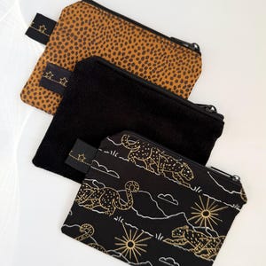 Può includere: Tre piccole pochette con cerniera. Una è color cammello con macchie marroni, una è in velluto a coste nero e la terza è nera con un disegno di leopardo e paesaggio dorato. Ogni pochette ha una linguetta nera con stelle dorate.