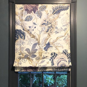 May include: A white roman shade with a floral pattern in shades of blue, purple, and gold. The shade is pulled up to reveal a window with a view of green foliage.