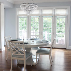 May include: A white dining table with six white chairs, a crystal chandelier, and four windows with white roman shades. The room is painted a light blue color.