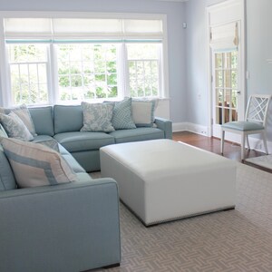 May include: A light blue sectional sofa with white and blue patterned pillows sits in a living room with a white ottoman and a patterned rug. The room has large windows with white blinds and a white door.