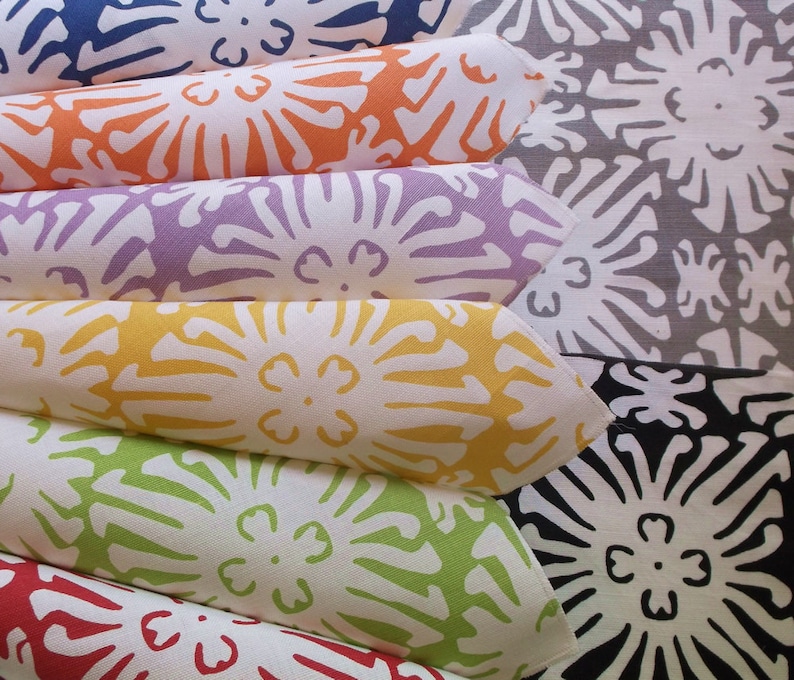 Puede incluir: Una pila de servilletas de tela dobladas en varios colores: azul, naranja, morado, amarillo, verde y rojo. Cada servilleta presenta un estampado floral blanco. El fondo muestra m&aacute;s servilletas en gris y negro.