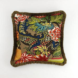 May include: A square throw pillow with a brown background and a colorful dragon design. The dragon is surrounded by flowers in shades of red, yellow, orange, and blue. The pillow has a brown fringe border.