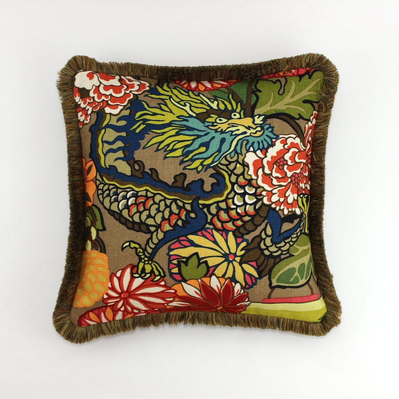 May include: A square throw pillow with a brown background and a colorful dragon design. The dragon is surrounded by flowers in shades of red, yellow, orange, and blue. The pillow has a brown fringe border.