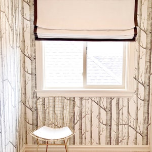 May include: A white roman shade with brown trim hangs in a window with a white frame. The window is in a room with white walls and a wallpaper pattern of birch trees. A gold wire chair sits in the corner of the room.