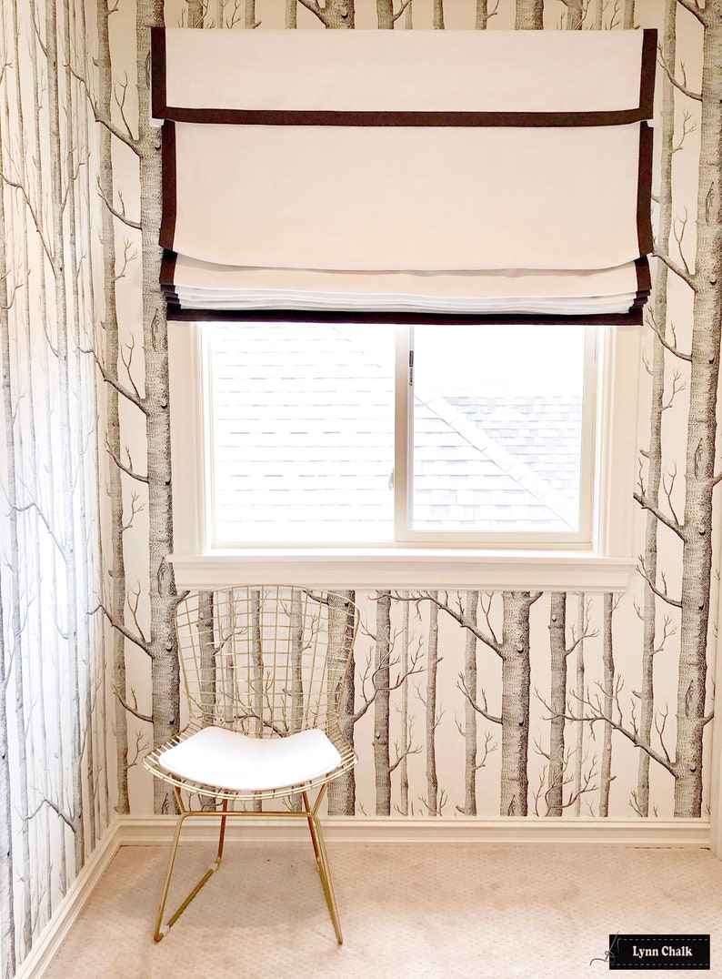 May include: A white roman shade with brown trim hangs in a window with a white frame. The window is in a room with white walls and a wallpaper pattern of birch trees. A gold wire chair sits in the corner of the room.