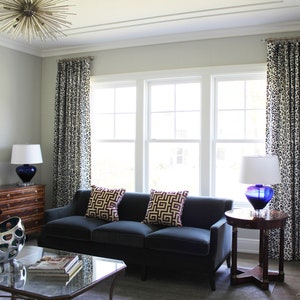 May include: A living room featuring a dark blue sofa, two throw cushions with geometric patterns, and a glass coffee table. The room has white walls, a large window with white trim, and a lamp with a blue glass base.