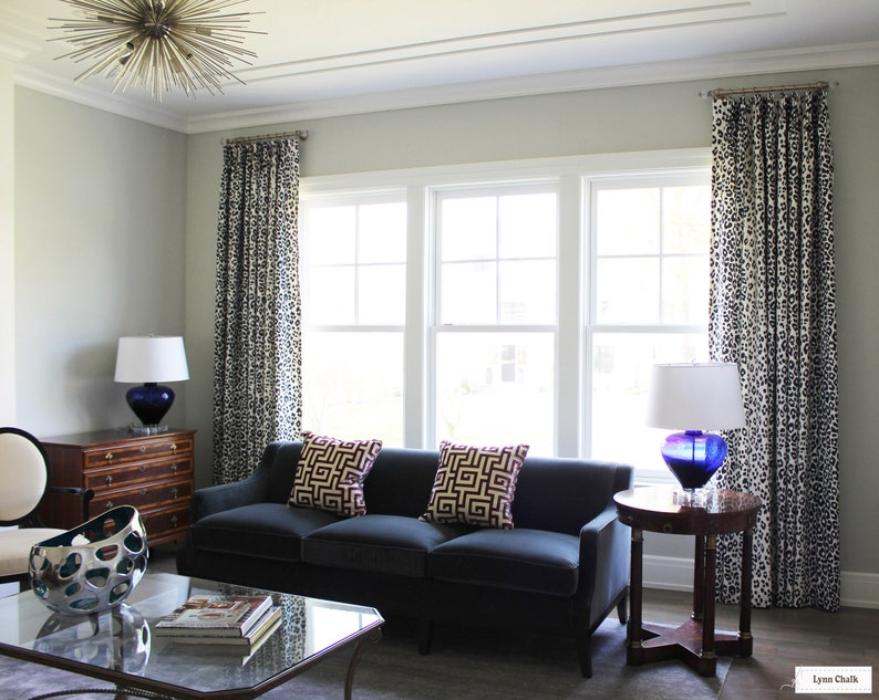 May include: A living room featuring a dark blue sofa, two throw cushions with geometric patterns, and a glass coffee table. The room has white walls, a large window with white trim, and a lamp with a blue glass base.