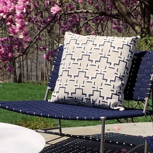 May include: A white outdoor chair with a navy blue woven seat and back. A white pillow with a navy blue geometric pattern is on the chair. The chair is on a patio with a pink magnolia tree in the background.
