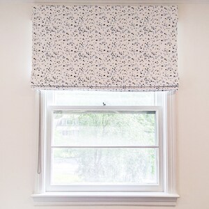 May include: White roman blind with a black and blue confetti pattern. The blind is pulled up to reveal a window with a view of green trees.