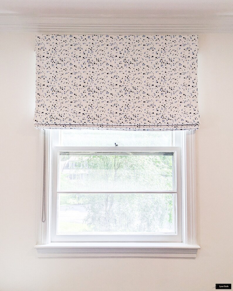 May include: White roman blind with a black and blue confetti pattern. The blind is pulled up to reveal a window with a view of green trees.