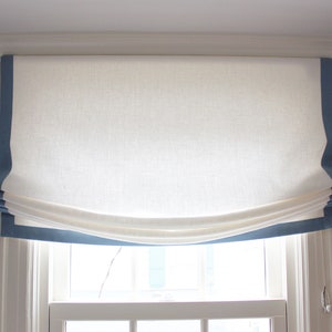 May include: White roman shade with a blue trim. The shade is pulled up and gathered in the center.