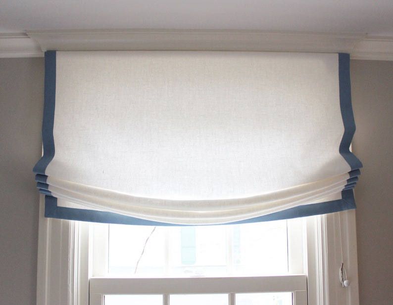 May include: White roman shade with a blue trim. The shade is pulled up and gathered in the center.