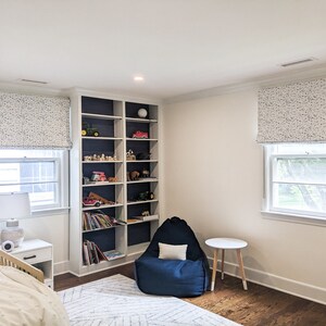 May include: A white bedroom with a bed, a bookshelf, a beanbag chair, and two windows with white roman blinds. The bookshelf is filled with toys and books. The beanbag chair is blue and has a white cushion. The windows have white trim and are covered with white roman blinds with a blue and white pattern.