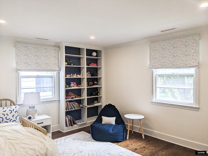 May include: A white bedroom with a bed, a bookshelf, a beanbag chair, and two windows with white roman blinds. The bookshelf is filled with toys and books. The beanbag chair is blue and has a white cushion. The windows have white trim and are covered with white roman blinds with a blue and white pattern.