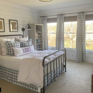 May include: A bedroom with a white bed with a blue and white checkered bed skirt and pillows. The bed has a black metal frame. There are two framed pictures on the wall behind the bed. There is a bookshelf to the right of the bed and a window with blue and white checkered curtains. The room has a light gray carpet.