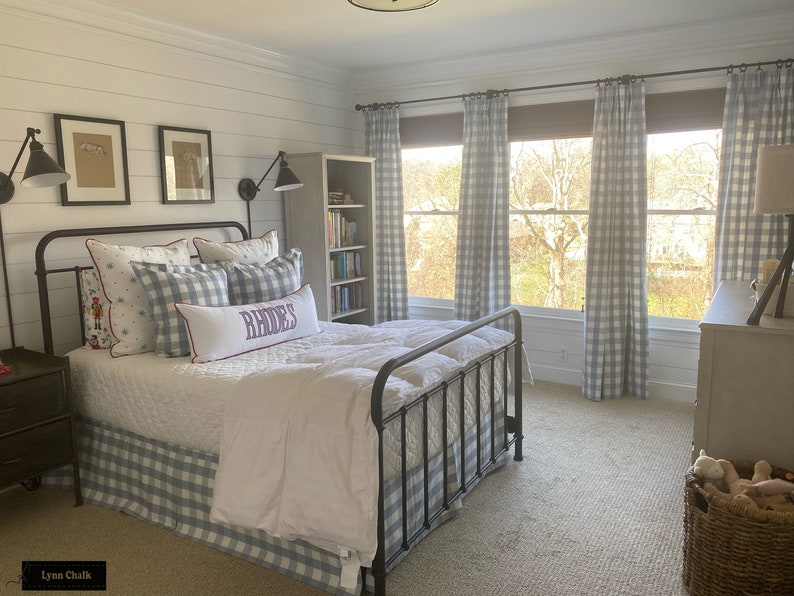 May include: A bedroom with a white bed with a blue and white checkered bed skirt and pillows. The bed has a black metal frame. There are two framed pictures on the wall behind the bed. There is a bookshelf to the right of the bed and a window with blue and white checkered curtains. The room has a light gray carpet.