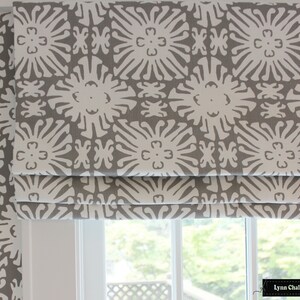 Puede incluir: Estores romanos estampados en gris y blanco. La tela presenta un dise&ntilde;o floral repetitivo en blanco sobre un fondo gris. Los estores est&aacute;n parcialmente bajados, revelando la ventana y la vista exterior.