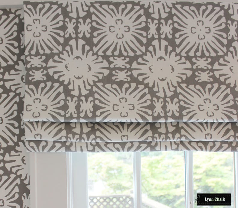 Puede incluir: Estores romanos estampados en gris y blanco. La tela presenta un dise&ntilde;o floral repetitivo en blanco sobre un fondo gris. Los estores est&aacute;n parcialmente bajados, revelando la ventana y la vista exterior.