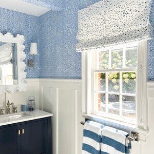 May include: A bathroom with white wainscoting and blue and white patterned wallpaper. A window with a white roman blind has a view of a green lawn. A dark blue vanity with a white countertop has a white porcelain sink and gold taps. Two blue and white striped towels hang on a chrome towel rail.