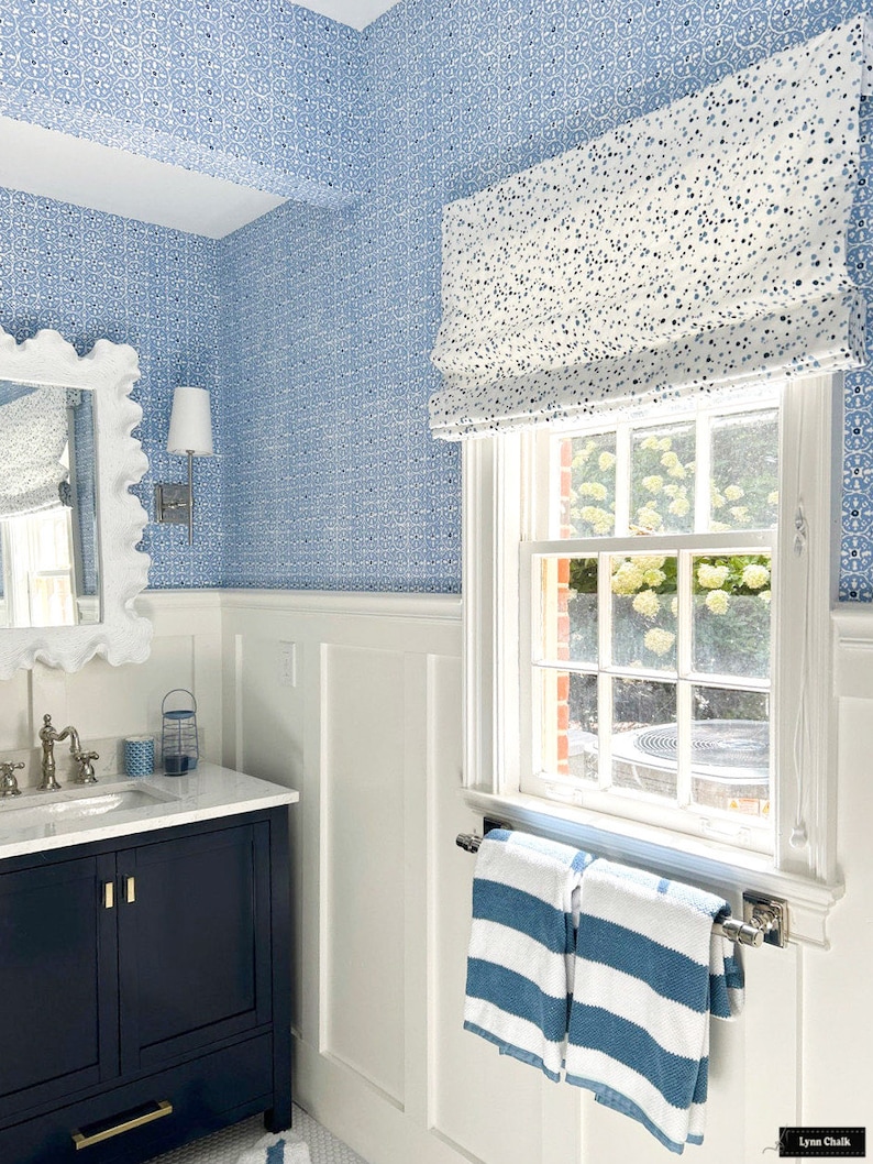 May include: A bathroom with white wainscoting and blue and white patterned wallpaper. A window with a white roman blind has a view of a green lawn. A dark blue vanity with a white countertop has a white porcelain sink and gold taps. Two blue and white striped towels hang on a chrome towel rail.